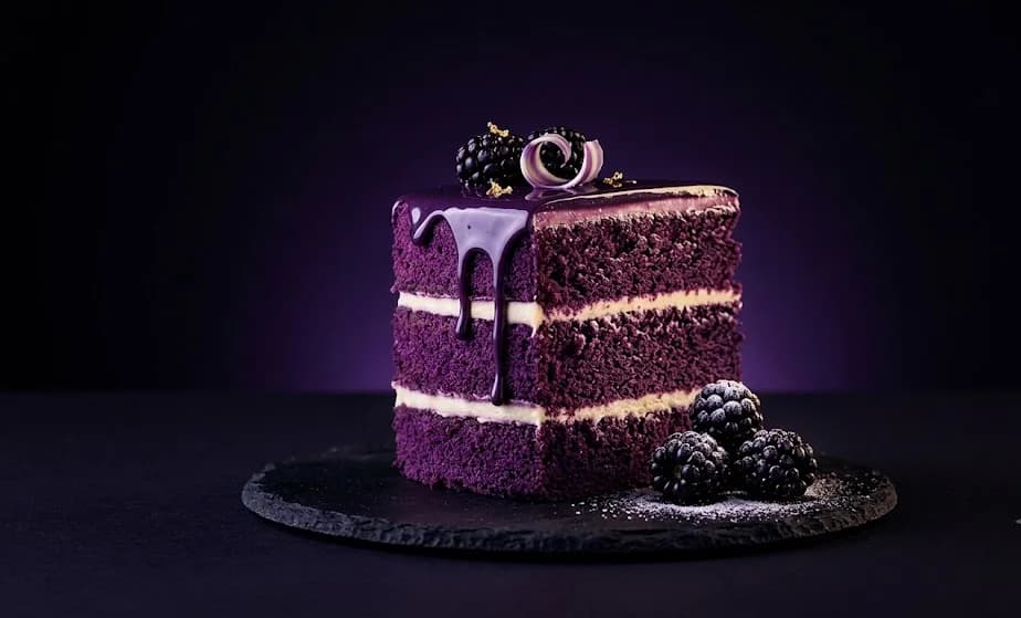 A tall slice of purple velvet layer cake with dark mirror glaze and sugared blackberries